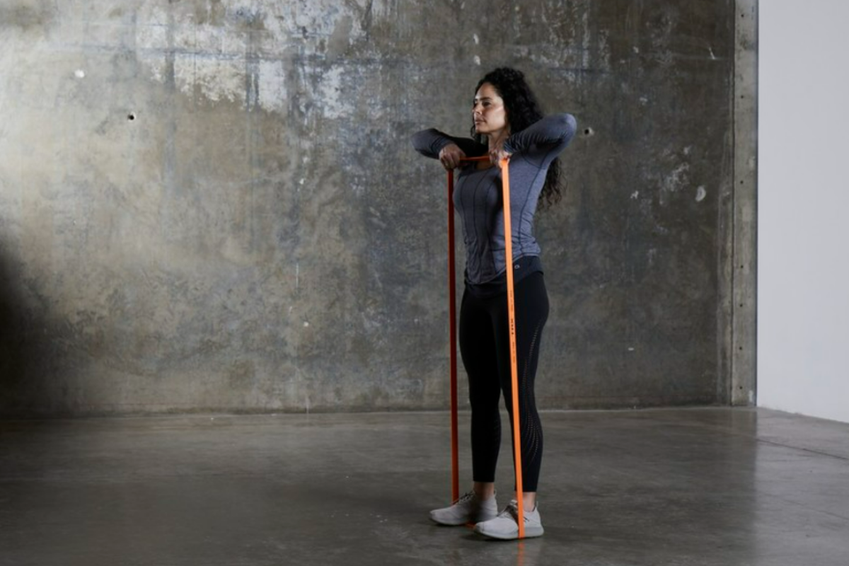 Woman in a minimalist studio stands on an orange resistance band and pulls it up toward her chest, demonstrating a tricep exercise with resistance bands.