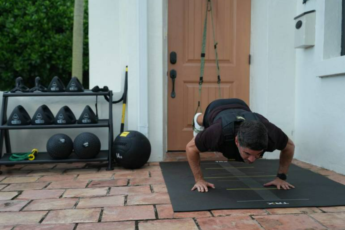 Man wearing a weighted vest does suspended push-ups on TRX straps on a patio beside kettlebells and medicine balls, demonstrating an at-home MMA workout.
