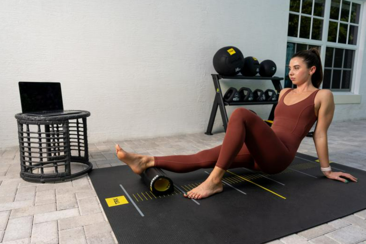 Woman in a rust-colored workout set uses a foam roller on her calf on a TRX mat next to a laptop and weight rack, illustrating careful, controlled exercise while recovering from injury.